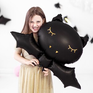 Girl holding cute black bat foil balloon with sleepy eyes – Halloween party kids decor, spooky bat balloon for children's Halloween celebration or themed birthday