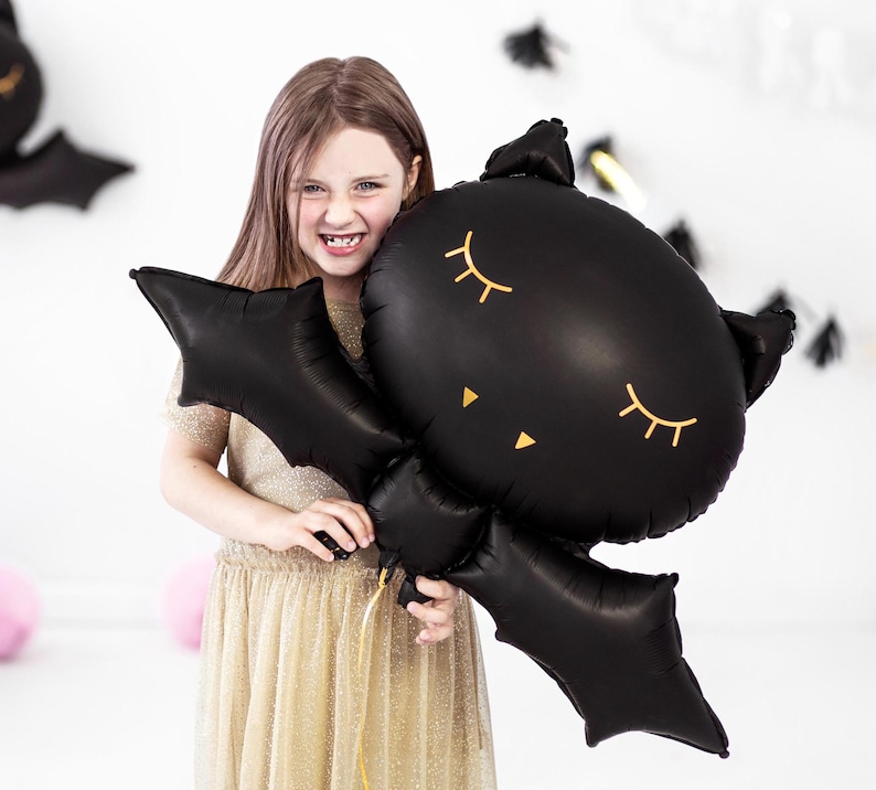 Girl holding cute black bat foil balloon with sleepy eyes – Halloween party kids decor, spooky bat balloon for children's Halloween celebration or themed birthday