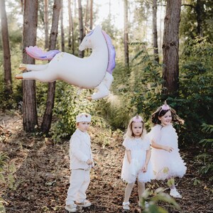 May include: Three children dressed in white and pink clothing stand in a woodland setting. A large white unicorn balloon with purple and gold accents floats above them. The children are wearing crowns and headbands.