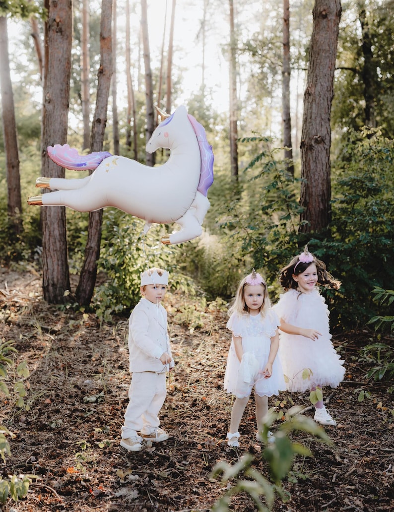May include: Three children dressed in white and pink clothing stand in a woodland setting. A large white unicorn balloon with purple and gold accents floats above them. The children are wearing crowns and headbands.