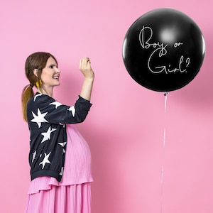 May include: A pregnant person in a pink dress and a black and white star patterned jacket is standing in front of a pink background. A black balloon with the text "Boy or Girl?" is floating in the air.