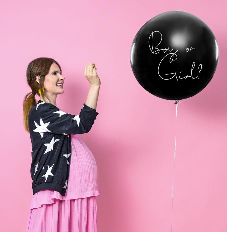 May include: A pregnant person in a pink dress and a black and white star patterned jacket is standing in front of a pink background. A black balloon with the text "Boy or Girl?" is floating in the air.