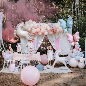 K&ouml;nnte beinhalten: Eine zauberhafte Geburtstagsfeier im Freien mit Einhorn-Thema. Die Dekoration umfasst pastellfarbene Ballons, Schmetterlingsakzente und ein Banner mit der Aufschrift "Happy Birthday". Kinder in Partykleidung sind anwesend, und ein rosa Raucheffekt verst&auml;rkt die magische Atmosph&auml;re.