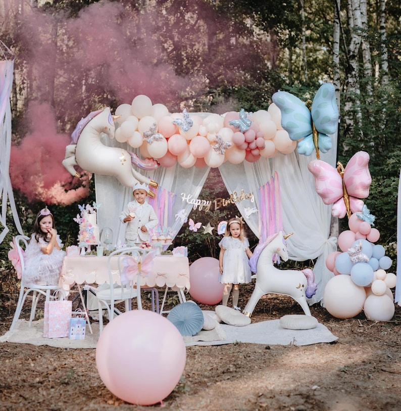 K&ouml;nnte beinhalten: Eine zauberhafte Geburtstagsfeier im Freien mit Einhorn-Thema. Die Dekoration umfasst pastellfarbene Ballons, Schmetterlingsakzente und ein Banner mit der Aufschrift "Happy Birthday". Kinder in Partykleidung sind anwesend, und ein rosa Raucheffekt verst&auml;rkt die magische Atmosph&auml;re.