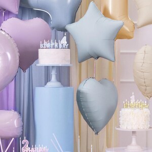May include: A festive scene with birthday cakes, balloons, and candles. A white cake with the word "BIRTHDAY" in silver candles sits on a pedestal. Pastel-colored heart and star-shaped balloons float in the background, creating a celebratory atmosphere.