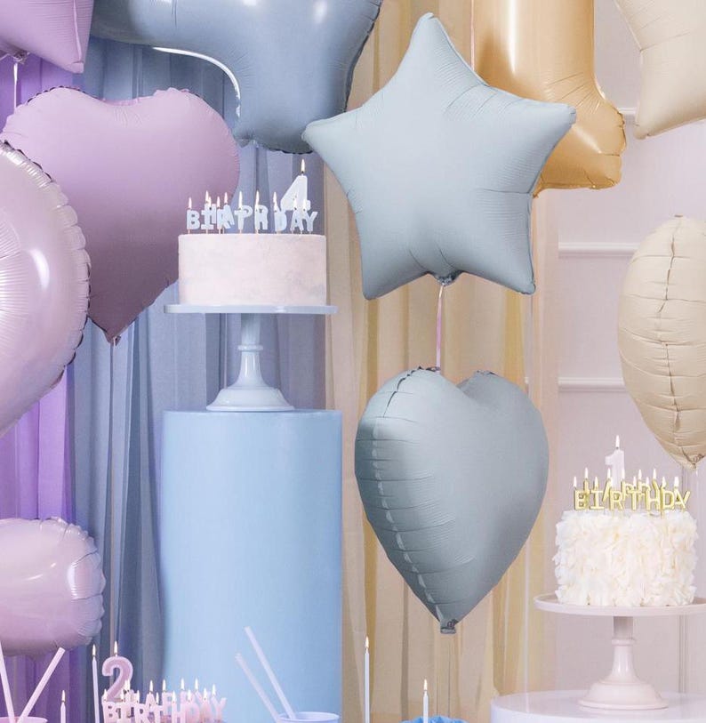 May include: A festive scene with birthday cakes, balloons, and candles. A white cake with the word "BIRTHDAY" in silver candles sits on a pedestal. Pastel-colored heart and star-shaped balloons float in the background, creating a celebratory atmosphere.