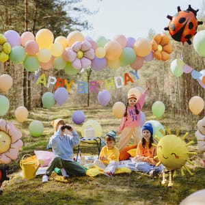 Outdoor children's birthday party with pastel balloons, flower and ladybug-shaped foil balloons, a 'Happy Birthday' banner, and kids playing and celebrating in a forest setting.