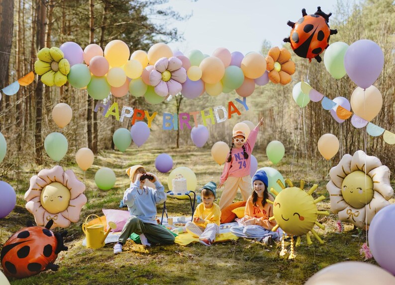 Outdoor children's birthday party with pastel balloons, flower and ladybug-shaped foil balloons, a 'Happy Birthday' banner, and kids playing and celebrating in a forest setting.