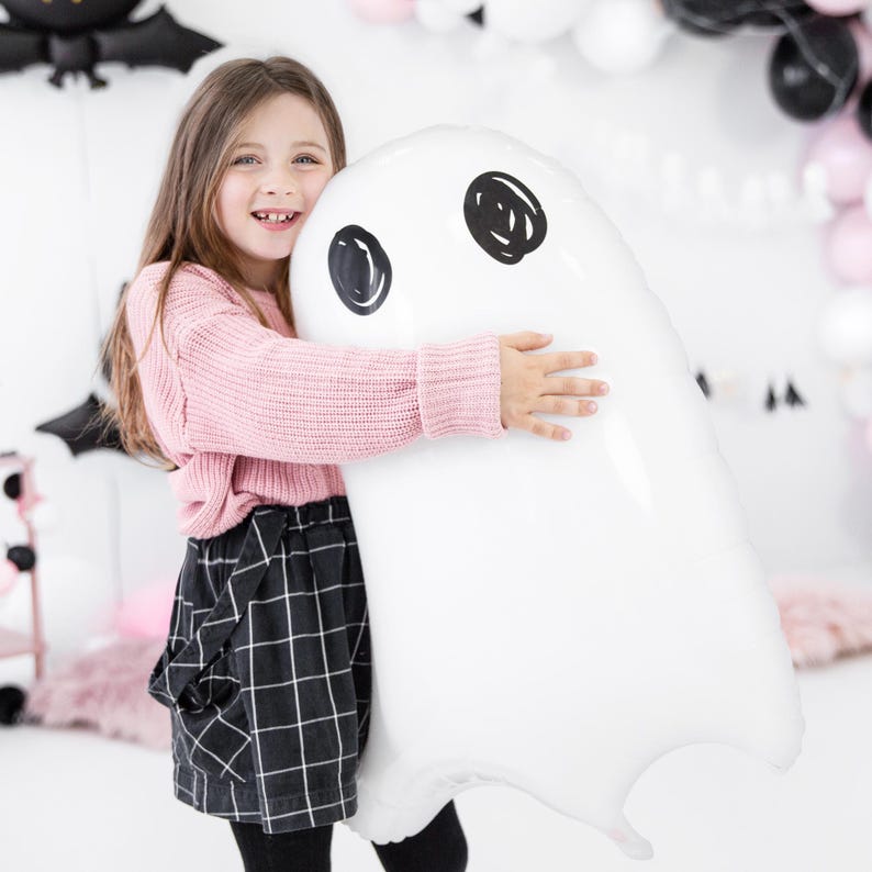 May include: A large white ghost-shaped balloon with black eye details is held by a person. The person is wearing a pink sweater and a black and white plaid skirt. The background includes black and pink balloons, suggesting a Halloween or party theme.