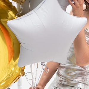 Op de afbeelding: Een zilveren stervormige ballon die door een persoon in een zilveren jurk wordt vastgehouden. De ballon is vastgebonden met witte linten.