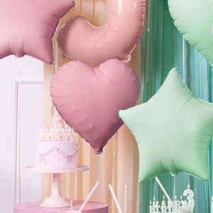 May include: A pink and green birthday party decoration set with balloons shaped like a star, heart, and a ring. The balloons are hanging from a ceiling or wall. There is a white cake with pink frosting and candles on a white cake stand.