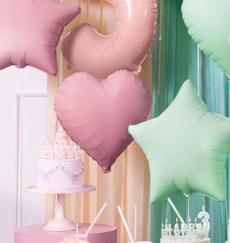 May include: A pink and green birthday party decoration set with balloons shaped like a star, heart, and a ring. The balloons are hanging from a ceiling or wall. There is a white cake with pink frosting and candles on a white cake stand.
