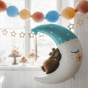 May include: A blue and white crescent moon balloon with gold stars and a brown bear balloon inside. The balloons are decorated with gold glitter and are hanging from a window.