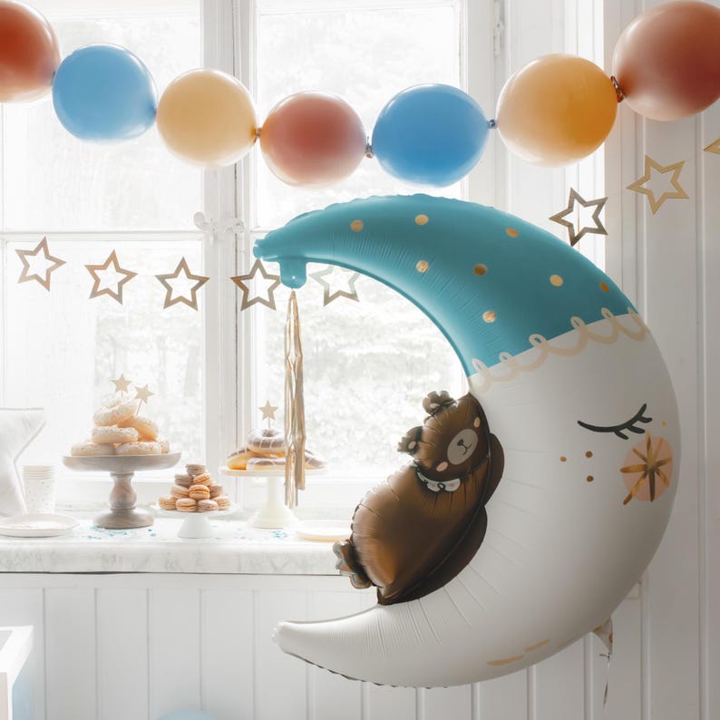 May include: A blue and white crescent moon balloon with gold stars and a brown bear balloon inside. The balloons are decorated with gold glitter and are hanging from a window.