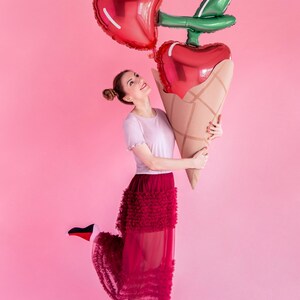 May include: A woman wearing a white shirt and a red tulle skirt holds two large heart-shaped balloons in the shape of a cherry. The balloons are red and green and are held in front of her. The background is pink.