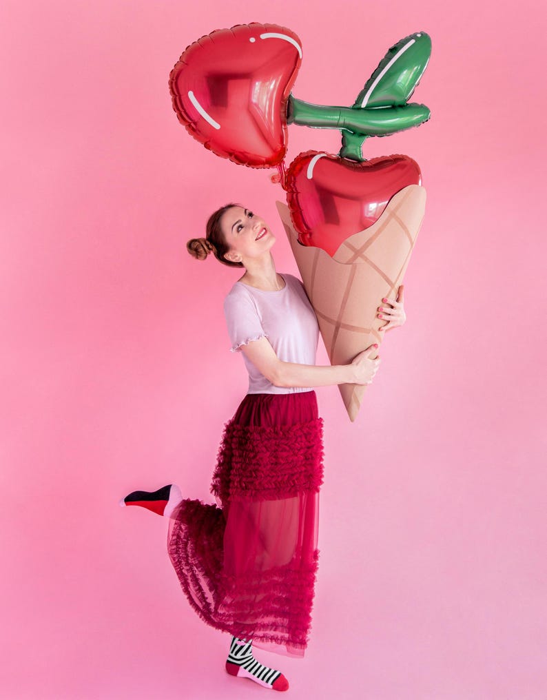 May include: A woman wearing a white shirt and a red tulle skirt holds two large heart-shaped balloons in the shape of a cherry. The balloons are red and green and are held in front of her. The background is pink.