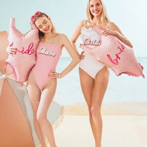 May include: Two women wearing pink and white swimsuits pose on a beach with pink foil balloons that say "Bride Babe" and "Bride".