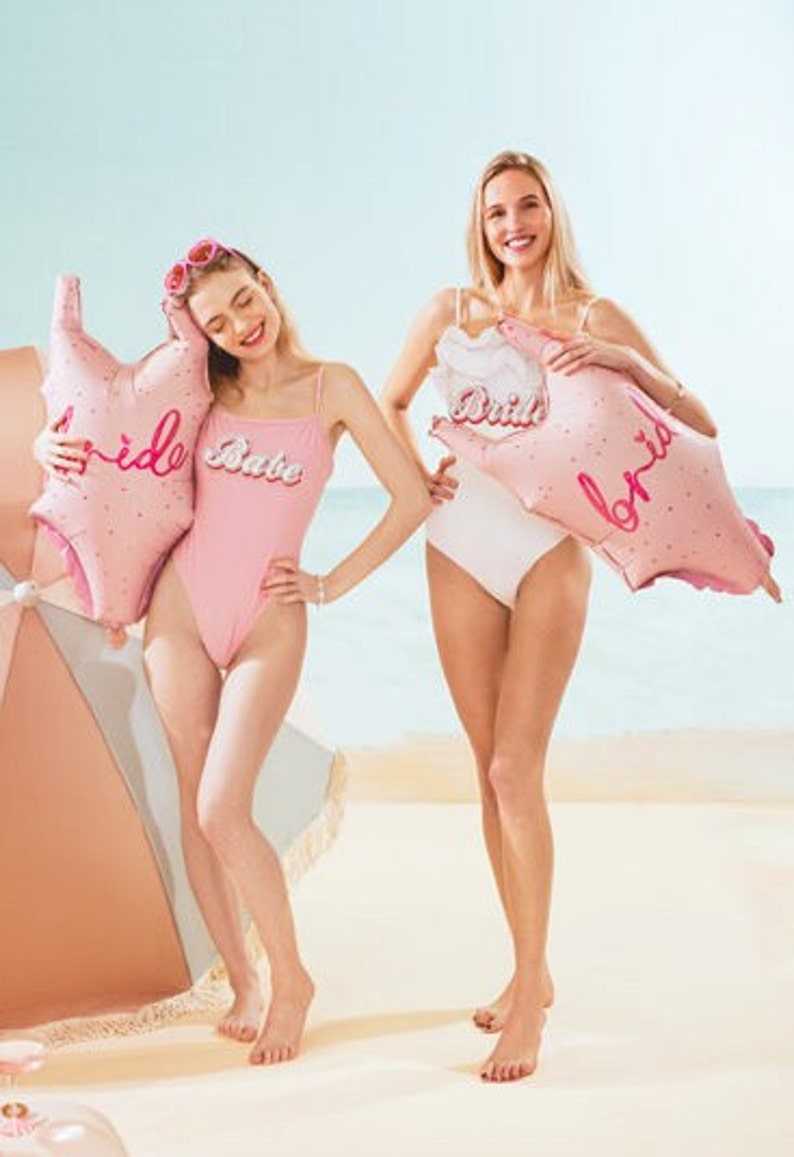 May include: Two women wearing pink and white swimsuits pose on a beach with pink foil balloons that say "Bride Babe" and "Bride".