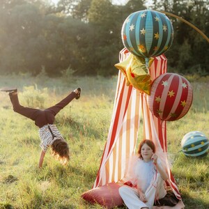 K&ouml;nnte beinhalten: Eine verspielte Szene im Freien mit einem rot-wei&szlig; gestreiften Zelt, bunten Luftballons und einem Kind, das einen Handstand macht. Die Luftballons sind mit Sternen und Streifen verziert. Ein zweites Kind sitzt im Zelt.