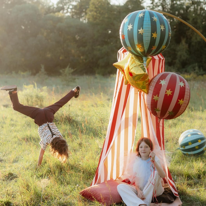 K&ouml;nnte beinhalten: Eine verspielte Szene im Freien mit einem rot-wei&szlig; gestreiften Zelt, bunten Luftballons und einem Kind, das einen Handstand macht. Die Luftballons sind mit Sternen und Streifen verziert. Ein zweites Kind sitzt im Zelt.