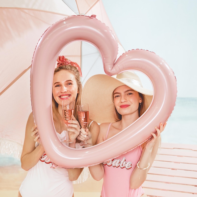 Light pink heart frame foil balloon held by two women at the beach, bachelorette party photo prop and bridal shower decor