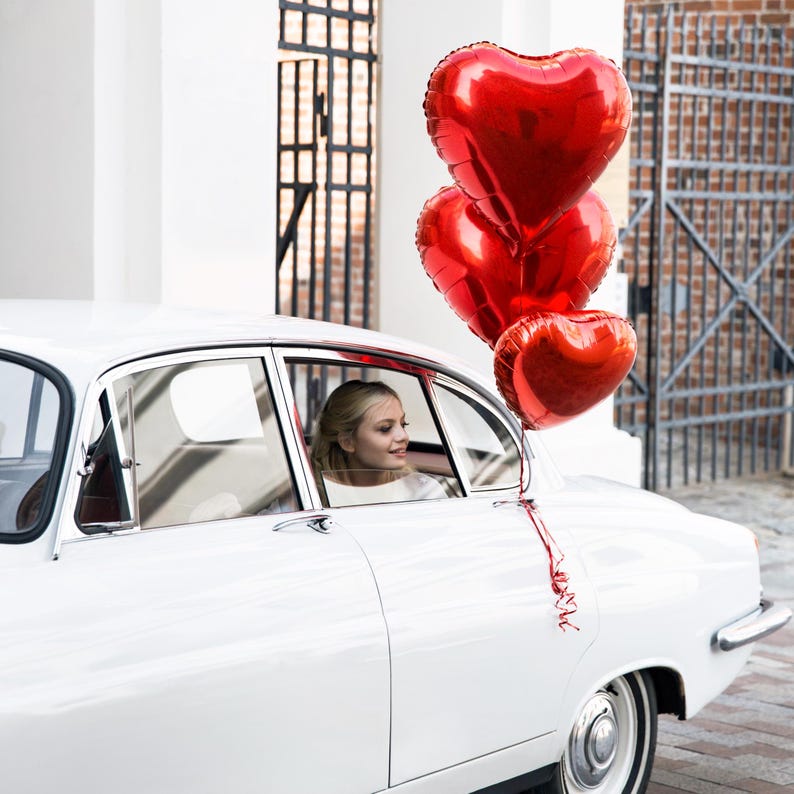 Puede incluir: Un coche vintage blanco con una mujer mirando por la ventana. Tres globos rojos en forma de coraz&oacute;n est&aacute;n atados a la antena del coche.