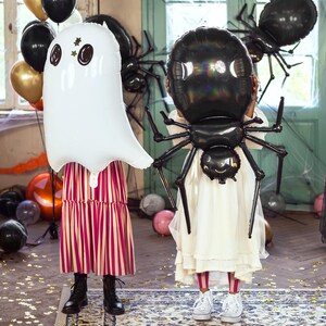 Op de afbeelding: Twee kinderen in witte jurken houden grote Halloween-ballonnen vast. E&eacute;n kind houdt een witte spookballon vast met zwarte ogen en een gouden ster. Het andere kind houdt een zwarte spinballon vast met een wit gezicht.