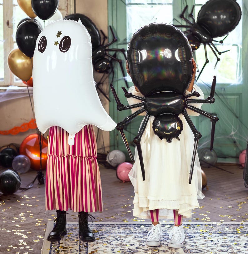 Op de afbeelding: Twee kinderen in witte jurken houden grote Halloween-ballonnen vast. E&eacute;n kind houdt een witte spookballon vast met zwarte ogen en een gouden ster. Het andere kind houdt een zwarte spinballon vast met een wit gezicht.
