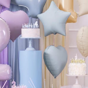 May include: A birthday party setup with a white cake with candles that spell out "birthday" and a blue star shaped balloon, a blue heart shaped balloon, and a light purple heart shaped balloon.
