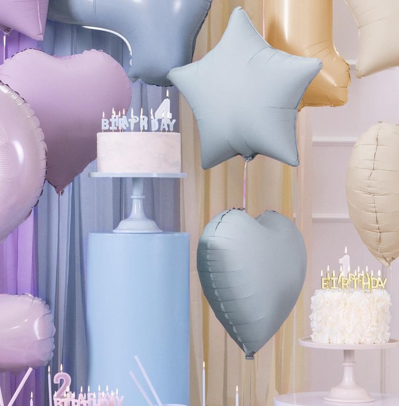 May include: A birthday party setup with a white cake with candles that spell out "birthday" and a blue star shaped balloon, a blue heart shaped balloon, and a light purple heart shaped balloon.