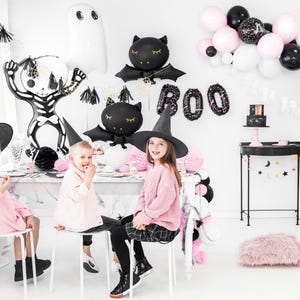 Halloween party setup for kids with pastel pink, black and white balloon decorations, including foil skeleton, ghost, and bat balloons by PartyDeco – spooky cute decor for children's Halloween celebration