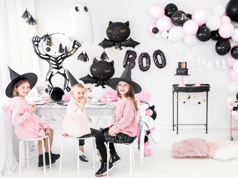 Halloween party setup for kids with pastel pink, black and white balloon decorations, including foil skeleton, ghost, and bat balloons by PartyDeco – spooky cute decor for children's Halloween celebration