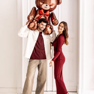 Pu&ograve; includere: Una coppia posa con un grande palloncino a forma di orsacchiotto di peluche marrone. L'uomo tiene il palloncino sopra la testa, mentre la donna sorride alla telecamera. Entrambi indossano abiti rossi.