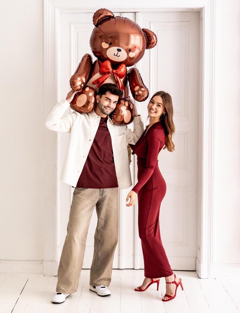 Pu&ograve; includere: Una coppia posa con un grande palloncino a forma di orsacchiotto di peluche marrone. L'uomo tiene il palloncino sopra la testa, mentre la donna sorride alla telecamera. Entrambi indossano abiti rossi.