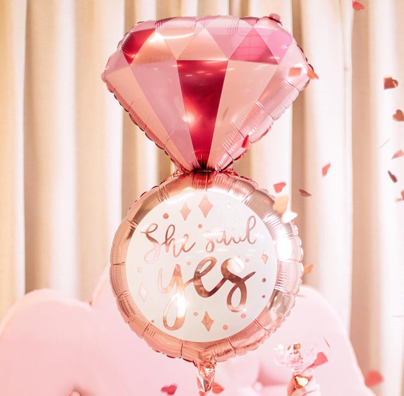 May include: A rose gold foil balloon shaped like a ring with a diamond on top. The ring balloon has the text "She said yes" on it.