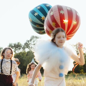 K&ouml;nnte beinhalten: Kinder rennen durch ein Feld, einige tragen Partykleidung. Zwei gro&szlig;e, bunte Ballons schweben &uuml;ber ihnen. Ein Ballon ist rot-wei&szlig; gestreift, der andere blau mit goldenen Sternen. Die Kinder sind auf einer Wiese mit B&auml;umen im Hintergrund.