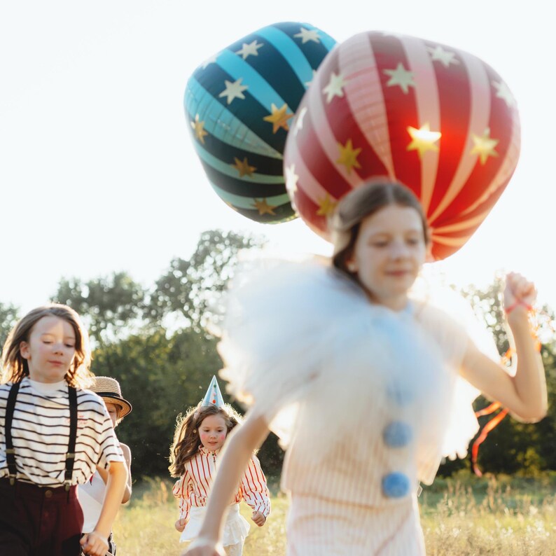 K&ouml;nnte beinhalten: Kinder rennen durch ein Feld, einige tragen Partykleidung. Zwei gro&szlig;e, bunte Ballons schweben &uuml;ber ihnen. Ein Ballon ist rot-wei&szlig; gestreift, der andere blau mit goldenen Sternen. Die Kinder sind auf einer Wiese mit B&auml;umen im Hintergrund.