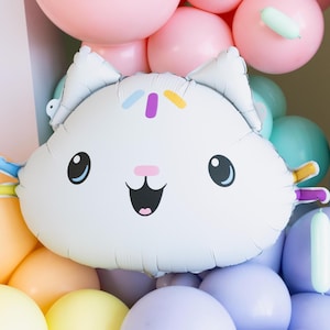 Könnte beinhalten: Ein weißer Ballon in Katzenform mit einem lächelnden Gesicht und bunten Streuseln. Der Ballon ist von einer Vielzahl pastellfarbener Ballons umgeben, darunter rosa, blau, gelb und lila, was eine festliche Atmosphäre schafft.