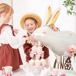 May include: Two children, one wearing a straw hat, are having a tea party with a white bunny balloon, cupcakes, and a teapot. The table is decorated with flowers and bunny ears.