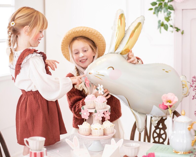 May include: Two children, one wearing a straw hat, are having a tea party with a white bunny balloon, cupcakes, and a teapot. The table is decorated with flowers and bunny ears.