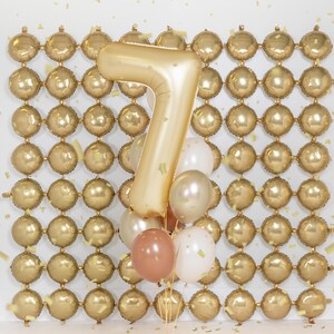 May include: A gold number seven balloon with a cluster of smaller balloons in white, gold, and brown against a backdrop of a gold balloon wall.