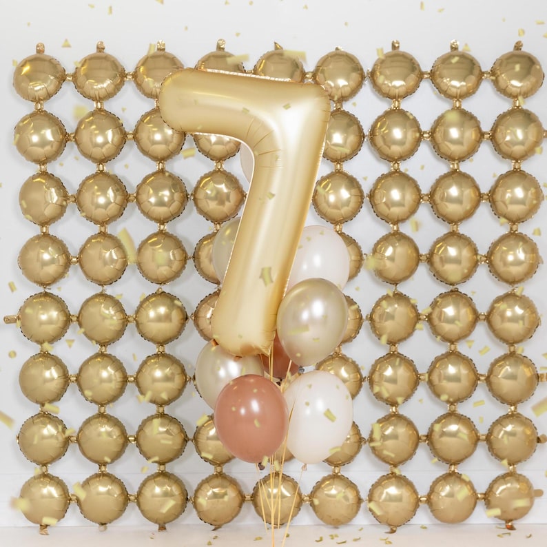 May include: A gold number seven balloon with a cluster of smaller balloons in white, gold, and brown against a backdrop of a gold balloon wall.