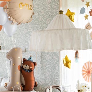 May include: A first birthday party table setting with a white canopy hanging overhead. The table is decorated with white flowers, candles, and a clear glass cake stand. There is a brown bear balloon and a gold number one balloon on the table. The background is a light green wallpaper with a floral pattern.