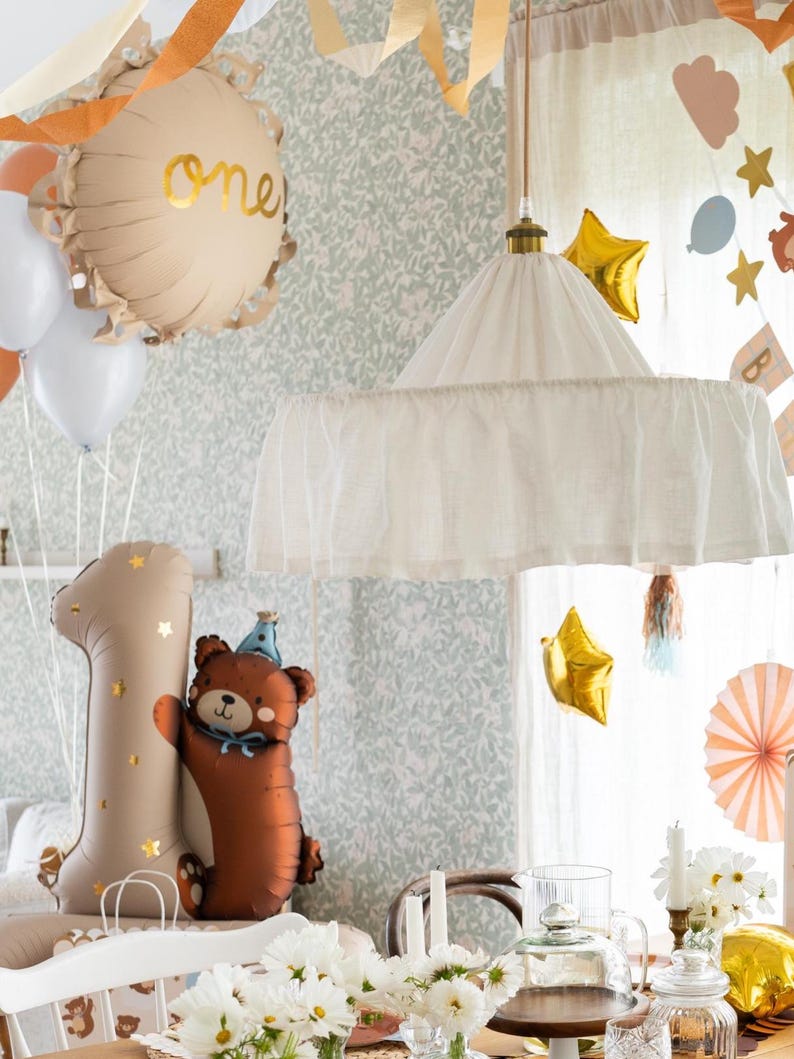 May include: A first birthday party table setting with a white canopy hanging overhead. The table is decorated with white flowers, candles, and a clear glass cake stand. There is a brown bear balloon and a gold number one balloon on the table. The background is a light green wallpaper with a floral pattern.