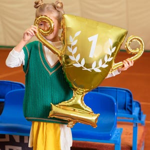 Puede incluir: Una persona joven con un chaleco de punto verde y una falda amarilla sostiene un globo de l&aacute;mina dorada con forma de trofeo. El trofeo tiene una corona de laurel blanca y el n&uacute;mero "1" en &eacute;l.