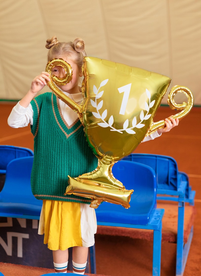 Puede incluir: Una persona joven con un chaleco de punto verde y una falda amarilla sostiene un globo de l&aacute;mina dorada con forma de trofeo. El trofeo tiene una corona de laurel blanca y el n&uacute;mero "1" en &eacute;l.