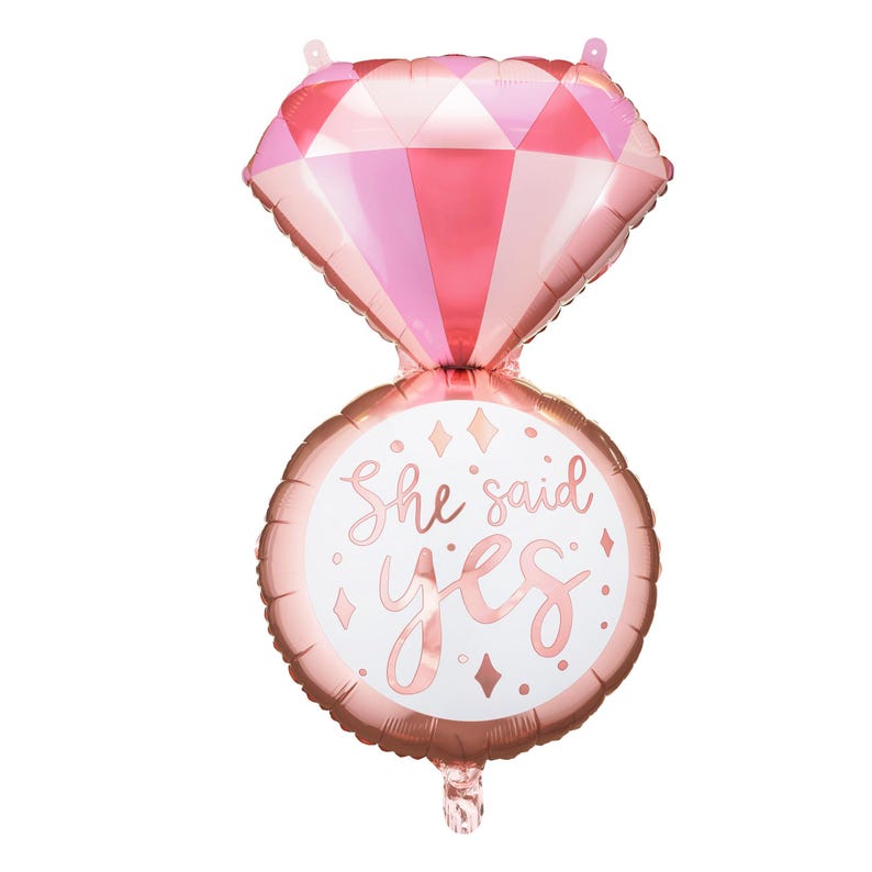 May include: A pink and rose gold foil balloon shaped like a ring with a diamond on top. The ring has the text "She said yes" on it.