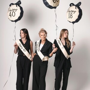 May include: Three women in black outfits holding balloons that say "Happy 40", "Happy 60", and "Happy 18". Each woman wears a sash with the same age on it. The balloons are cream-colored with black trim and bows.