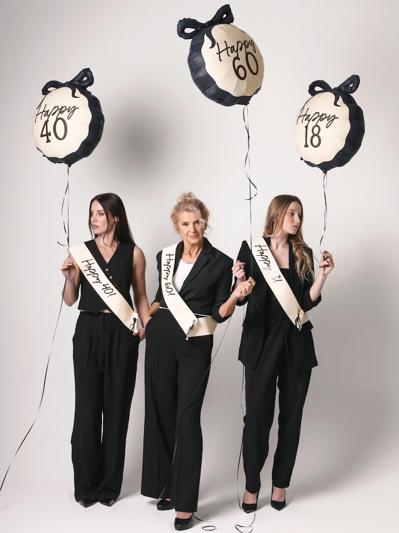 May include: Three women in black outfits holding balloons that say "Happy 40", "Happy 60", and "Happy 18". Each woman wears a sash with the same age on it. The balloons are cream-colored with black trim and bows.