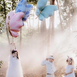May include: Three children dressed in white clothing and wearing unicorn and crown headbands play in a foggy forest with three large butterfly balloons. The balloons are pink, blue, and blue with white polka dots.
